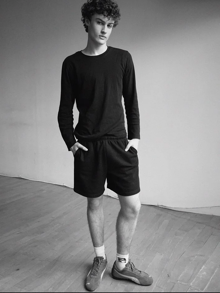 A young person with curly hair standing in a room with wooden floors and a plain wall, wearing a black long-sleeve shirt, black shorts, and sneakers.