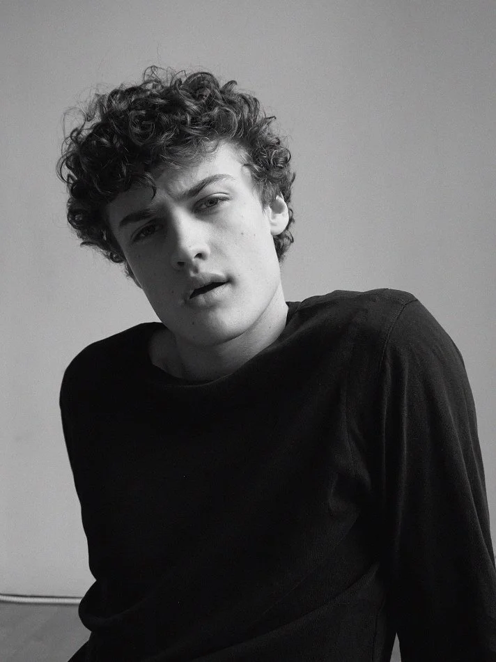 Albert, Montréal male model signed with MTL Models MGMT with curly hair and a serious expression, modelling at the camera, wearing a black shirt, in a black and white photo against a plain background.