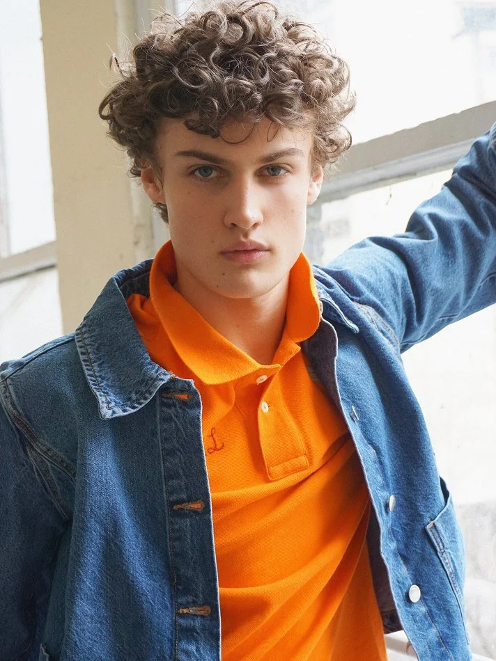 Albert, Montréal male model signed with MTL Models MGMT with curly hair wearing an orange polo shirt and a denim jacket, standing indoors near a window.