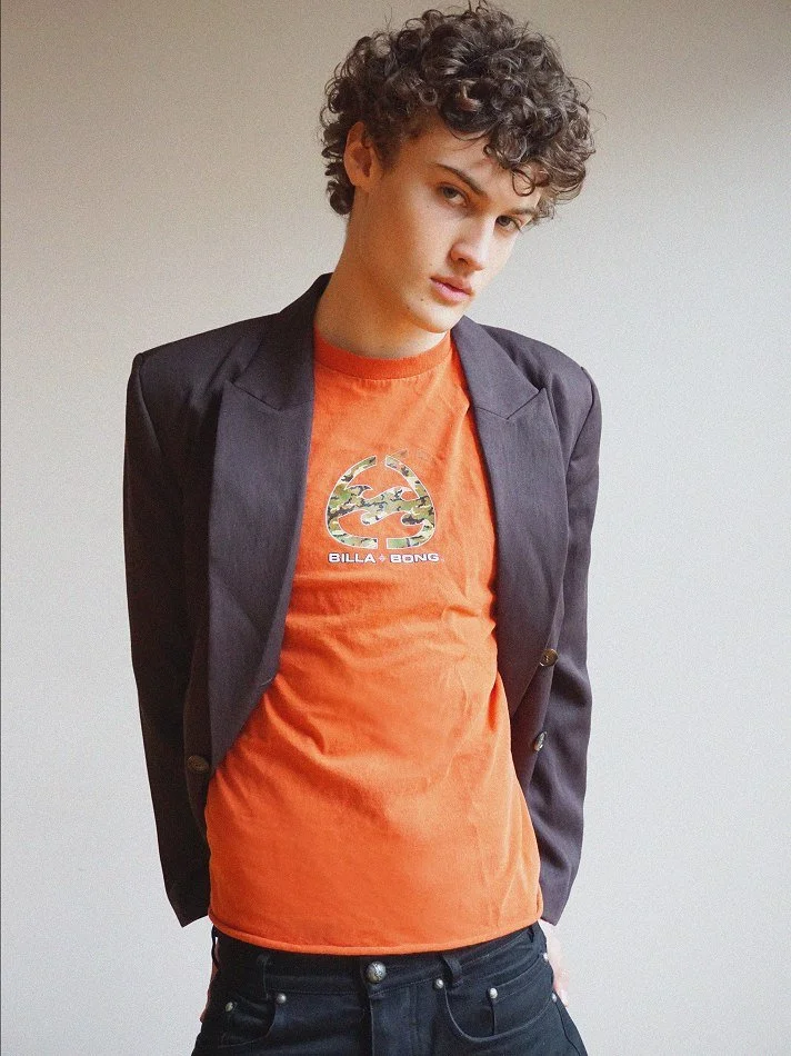 A young man with curly dark hair wearing an orange t-shirt with a graphic logo and text, paired with a dark blazer, standing against a plain light-colored wall.