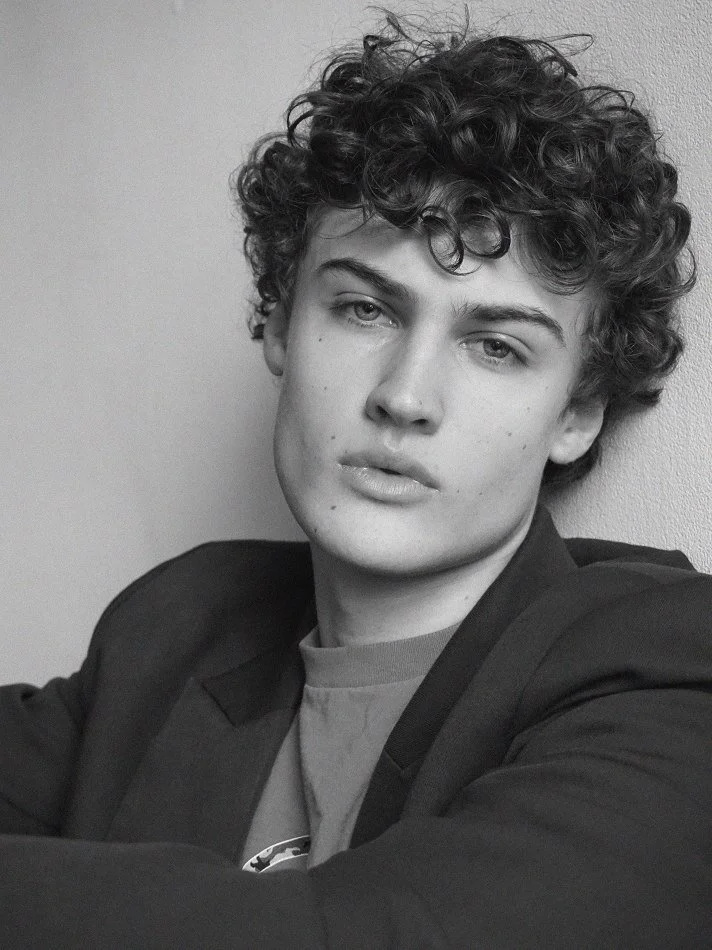 Albert, Montréal male model signed with MTL Models MGMT with curly hair and light skin, wearing a dark blazer over a T-shirt, looking at the camera with a neutral expression.