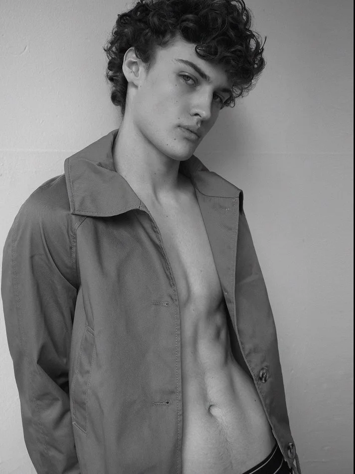 Black and white photo of a young man with curly hair, wearing an open jacket exposing his bare chest and abdomen, standing against a plain wall.
