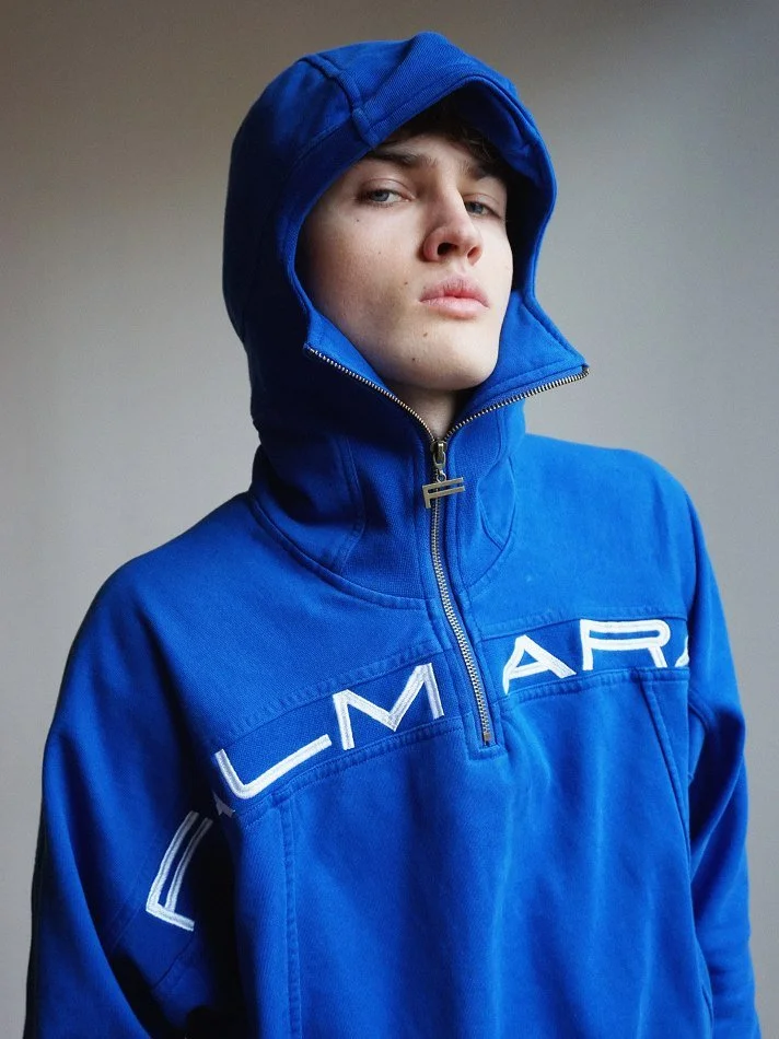 Young man wearing a blue hooded jacket with the word 'LMAIR' on the front, looking confidently at the camera.