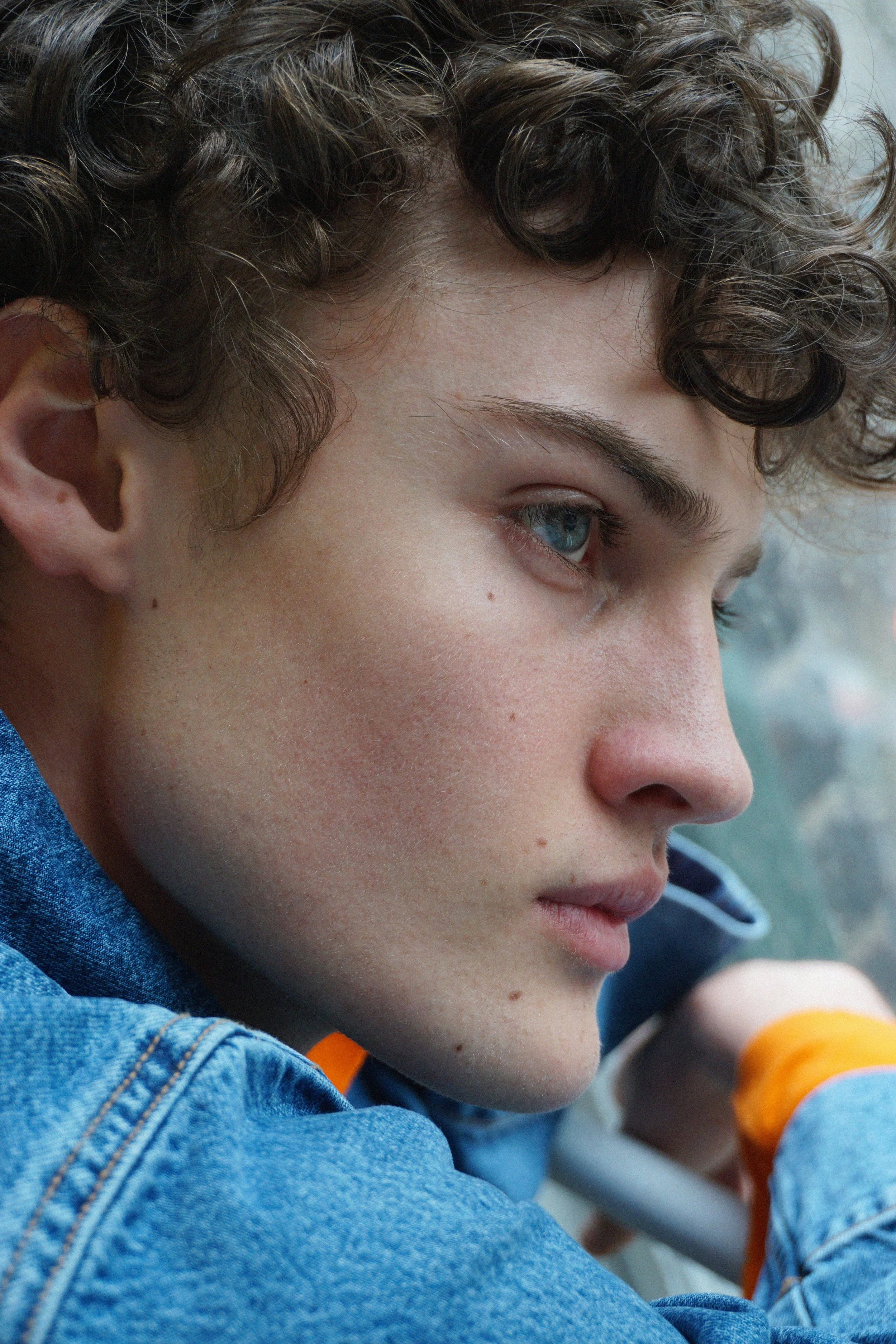 Close-up of a young boy with curly brown hair and blue eyes, wearing a denim jacket, sitting outdoors, with a blurred background.