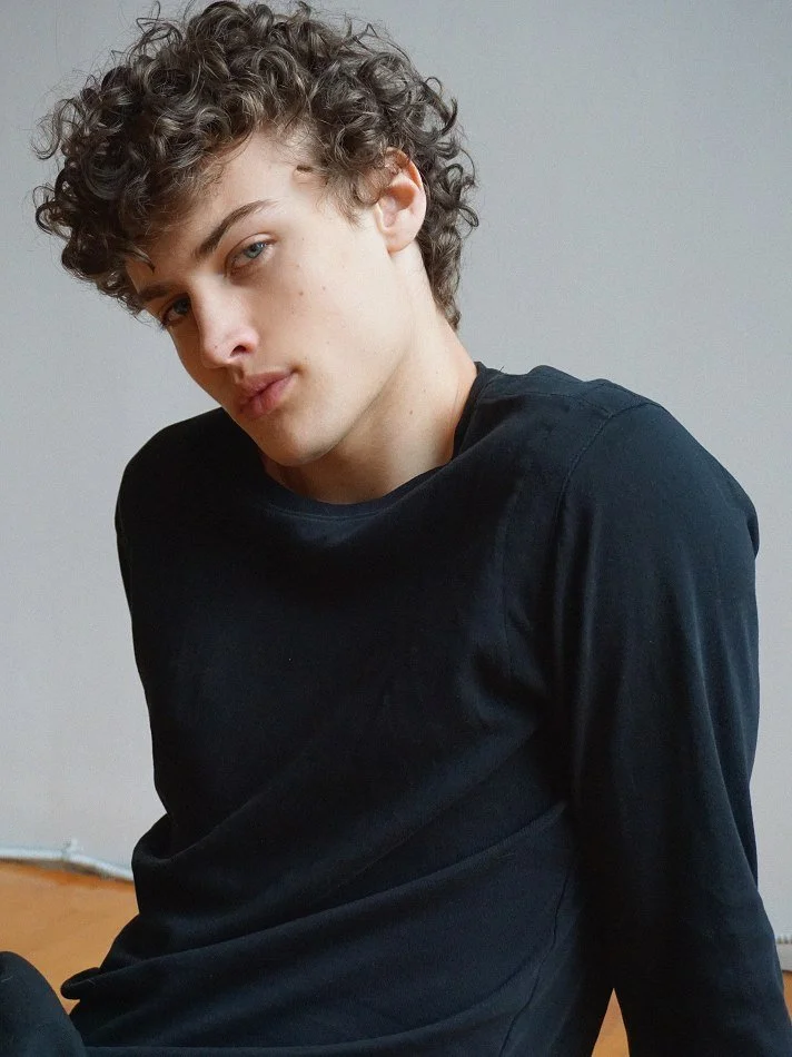 Albert, Montréal male model signed with MTL Models MGMT with curly hair and fair skin wearing a black shirt, looking at the camera with a serious expression, sitting in an indoor setting with a plain background.