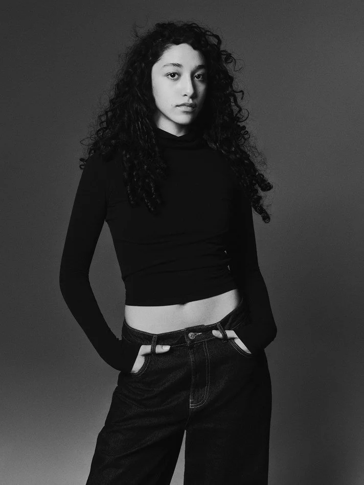 Black and white portrait of Oana, Montréal male model signed with MTL Models MGMT with curly hair, wearing a long-sleeved black crop top and high-waisted jeans, posing against a plain background in a Montreal photo studio.