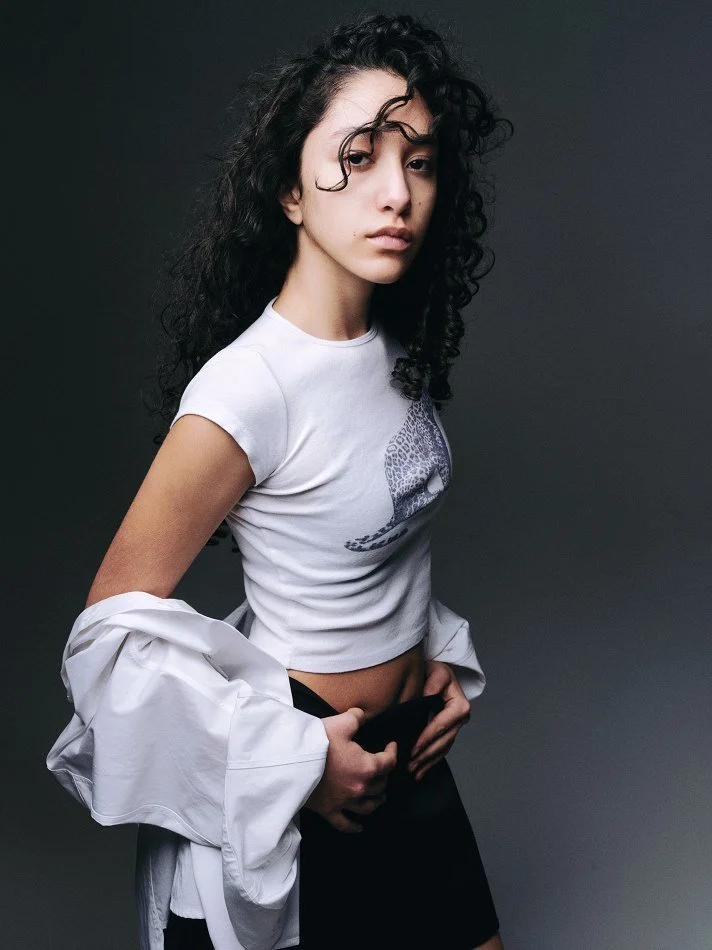 Oana, Montréal male model signed with MTL Models MGMT with curly black hair, wearing a white crop top with a cheetah print design, partially removed a white jacket revealing black shorts, against a dark gray background.