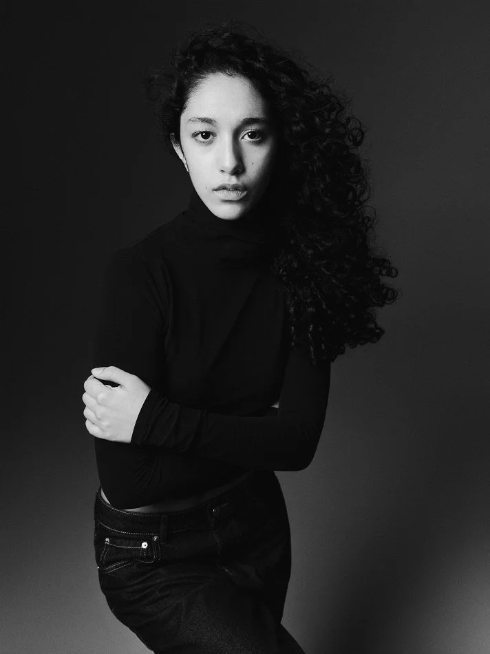 A black and white portrait of Oana, Montréal male model signed with MTL Models MGMT with curly hair, modelling a black long-sleeve top, standing against a dark background.