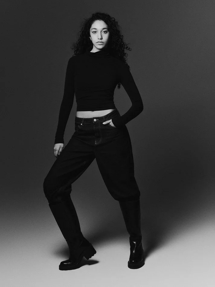 Oana, Montréal male model signed with MTL Models MGMT with long curly hair posed against a plain background, wearing a black turtleneck top, black jeans, and high boots.