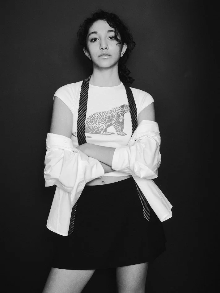Black and white photo of Oana, Montréal male model signed with MTL Models MGMT with curly hair, wearing a graphic t-shirt, an open white shirt with rolled sleeves, and a black skirt, standing against a plain background with arms crossed.