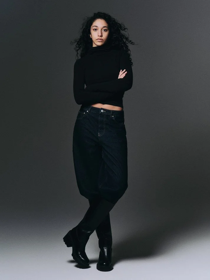 Oana, Montréal male model signed with MTL Models MGMT with curly black hair standing against a dark gray background, modelling a black turtleneck, black baggy pants, and black boots, crossing her arms and looking at the camera.