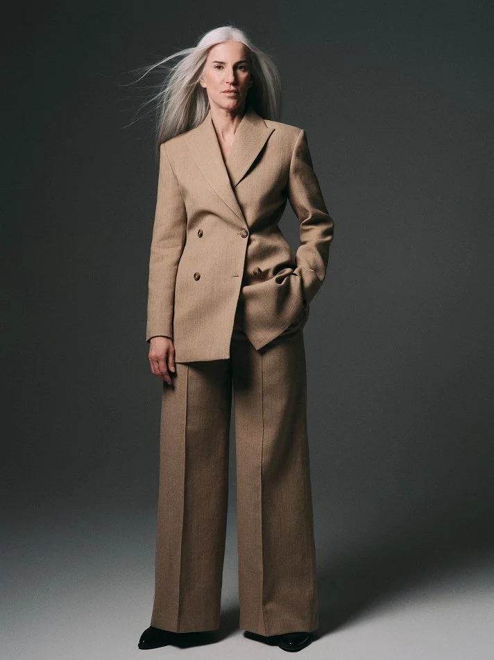 Carmen, Montréal female mature model signed with MTL Models MGMT with long gray hair is wearing a beige double-breasted blazer and matching wide-leg trousers, standing against a dark gray background.