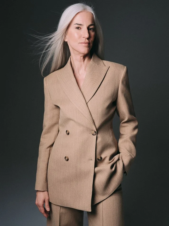 Carmen, Montréal female mature model signed with MTL Models MGMT with long silver hair wearing a tan double-breasted blazer and matching pants against a dark gray background.