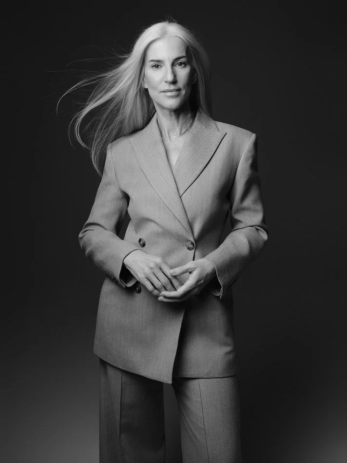 Black and white photograph of Carmen, Montréal female mature model signed with MTL Models MGMT with long, flowing silver hair wearing a tailored suit jacket and matching trousers, standing against a dark background.