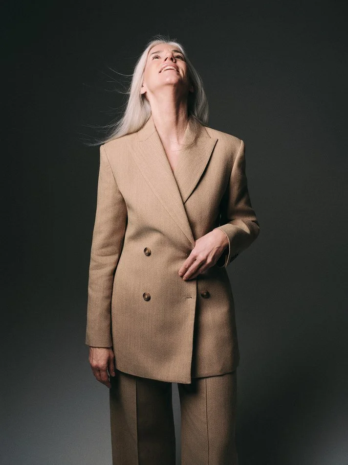 An older female model, Carmen, Montréal female mature model signed with MTL Models MGMT with long gray hair wearing a tan blazer and matching pants standing against a dark background, looking up with a slight smile.