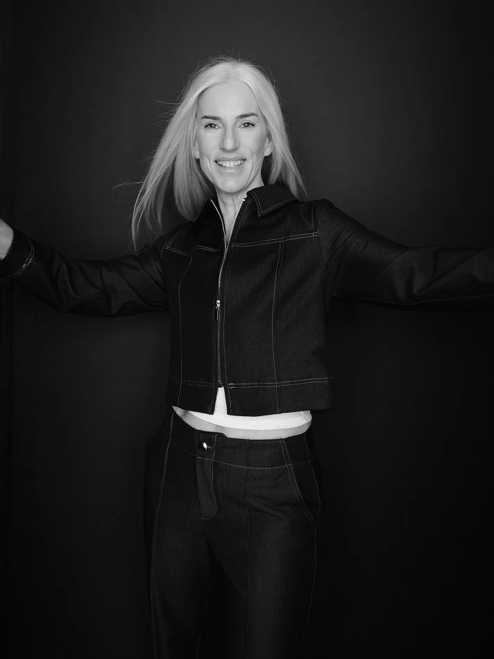 Smiling Carmen, Montréal female mature model signed with MTL Models MGMT with long light hair wearing a black jacket and pants against a dark background.