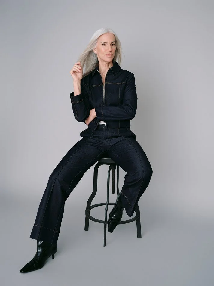 A stylish older Carmen, Montréal female model signed with MTL Models MGMT  with long gray hair, wearing a dark denim jacket and matching wide-leg jeans, sitting on a black stool against a plain gray background.