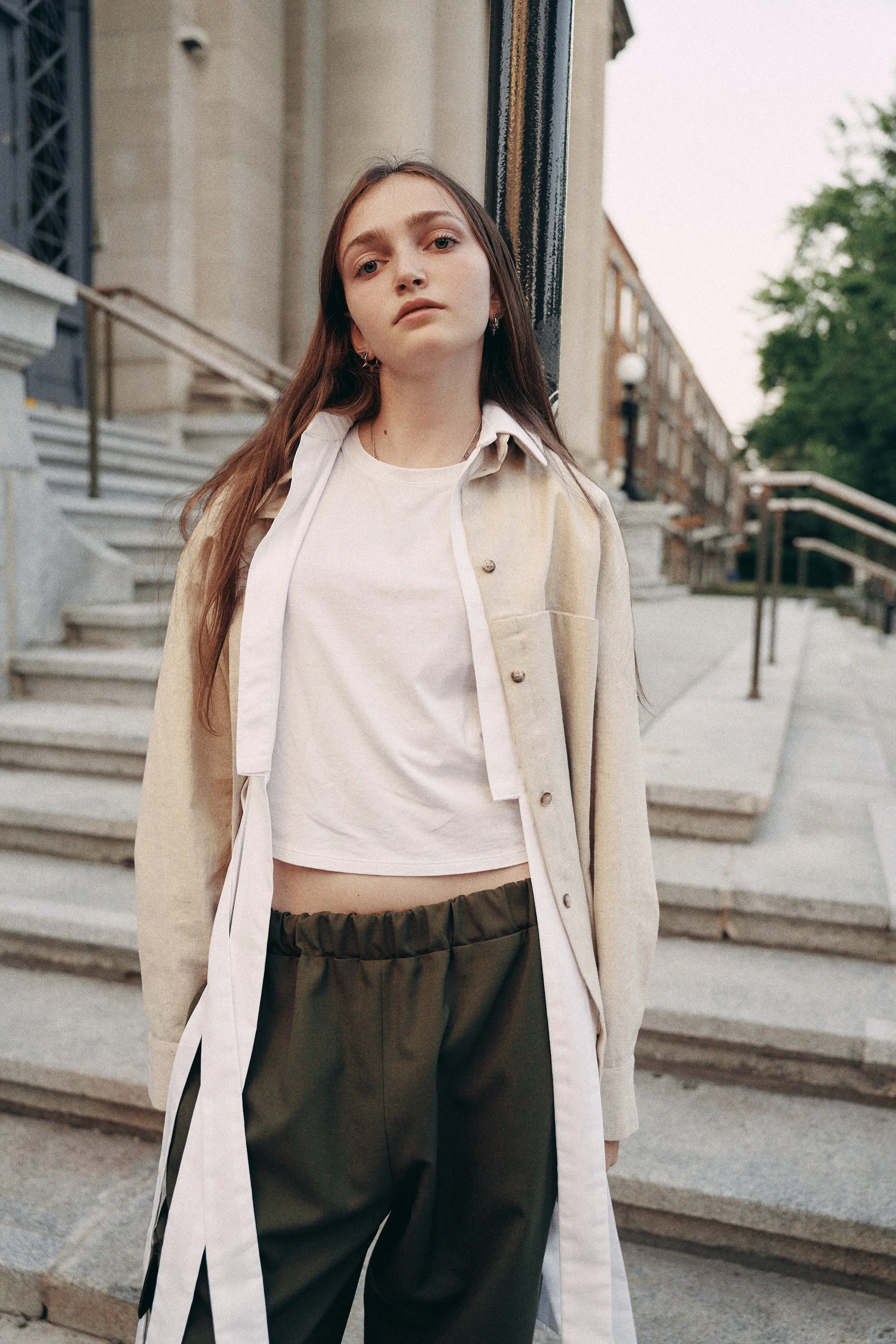 A young woman with long brown hair leaning against a black pole outdoors. She is wearing a beige coat over a white T-shirt and dark green pants. The background features concrete steps, a building with beige and brown tones, and some trees, indicating