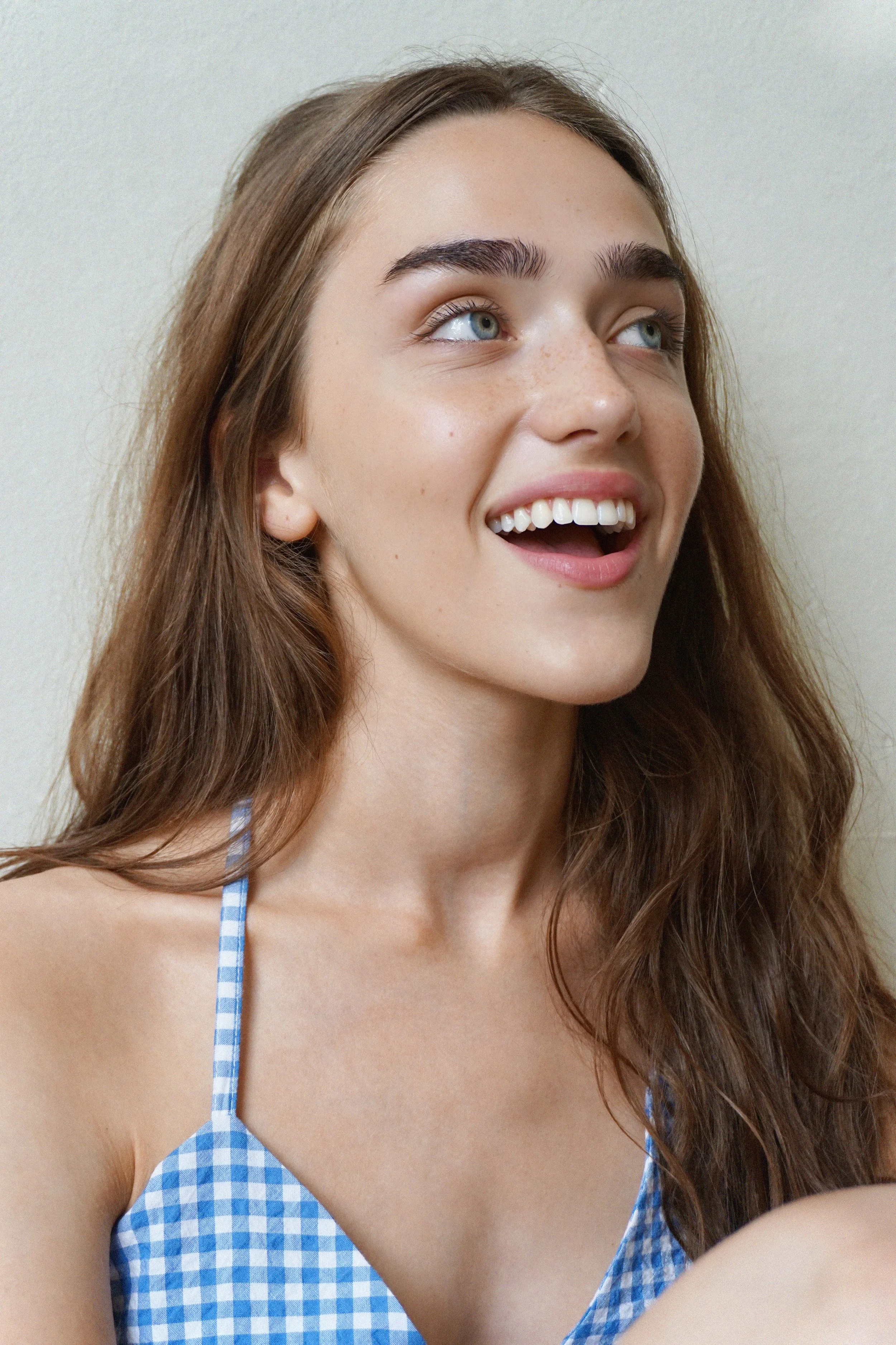 Close-up of a young woman with long brown hair wearing a blue and white checkered spaghetti strap top, smiling with her teeth visible and looking upward.
