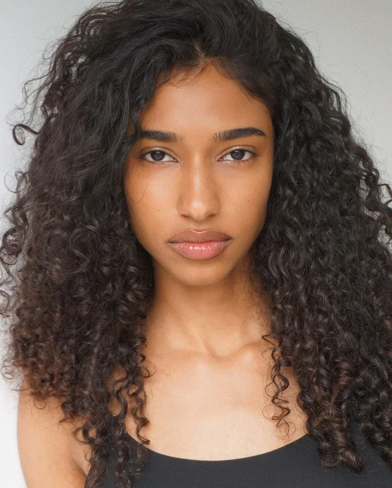 A young woman with long, dark, curly hair and a neutral expression, wearing a black top, looking directly at the camera against a plain background.