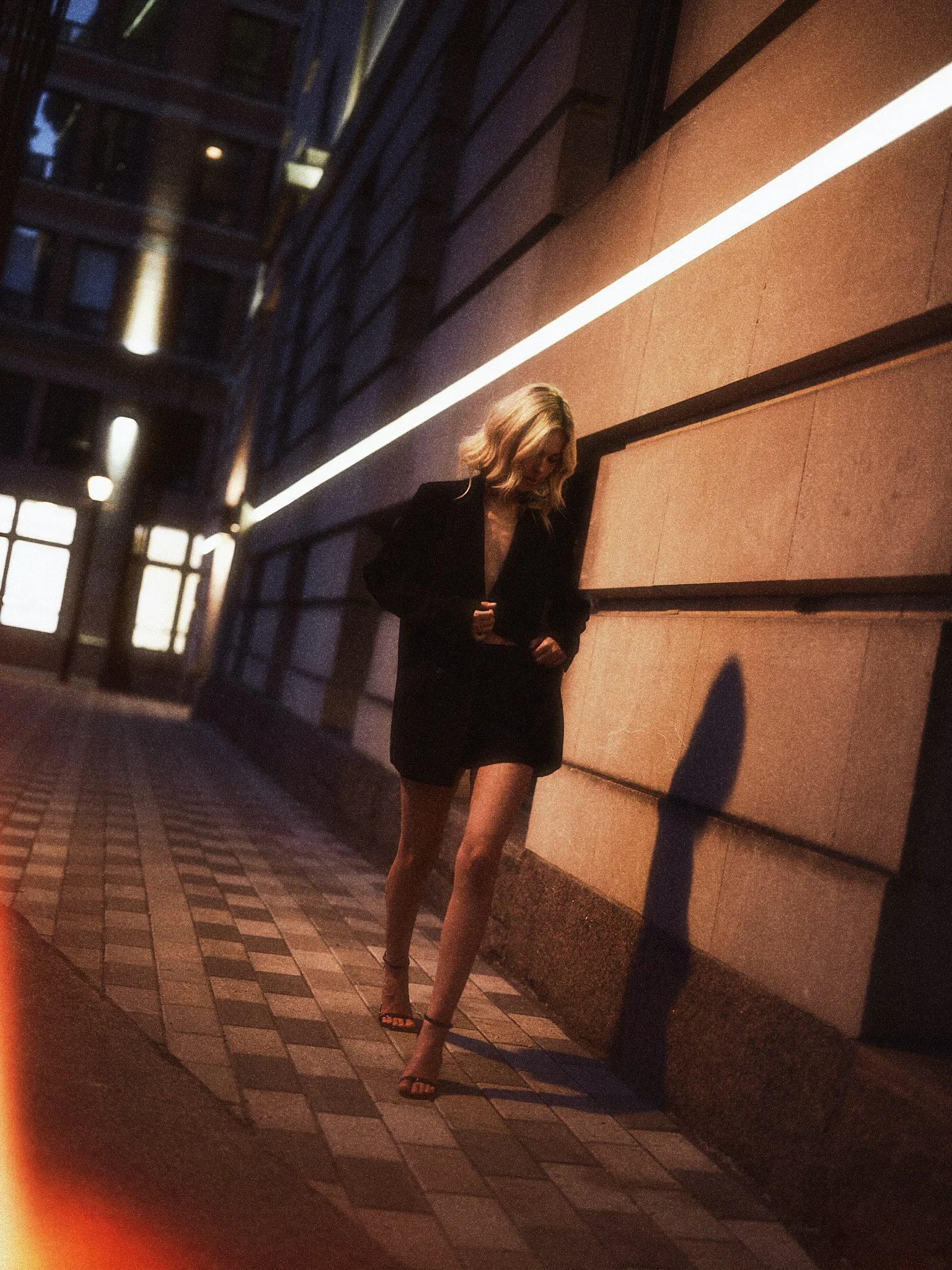 A woman with blonde hair in a black dress and high heels walking outside at night near a building with a brick and stone facade, illuminated by a bright white light strip.