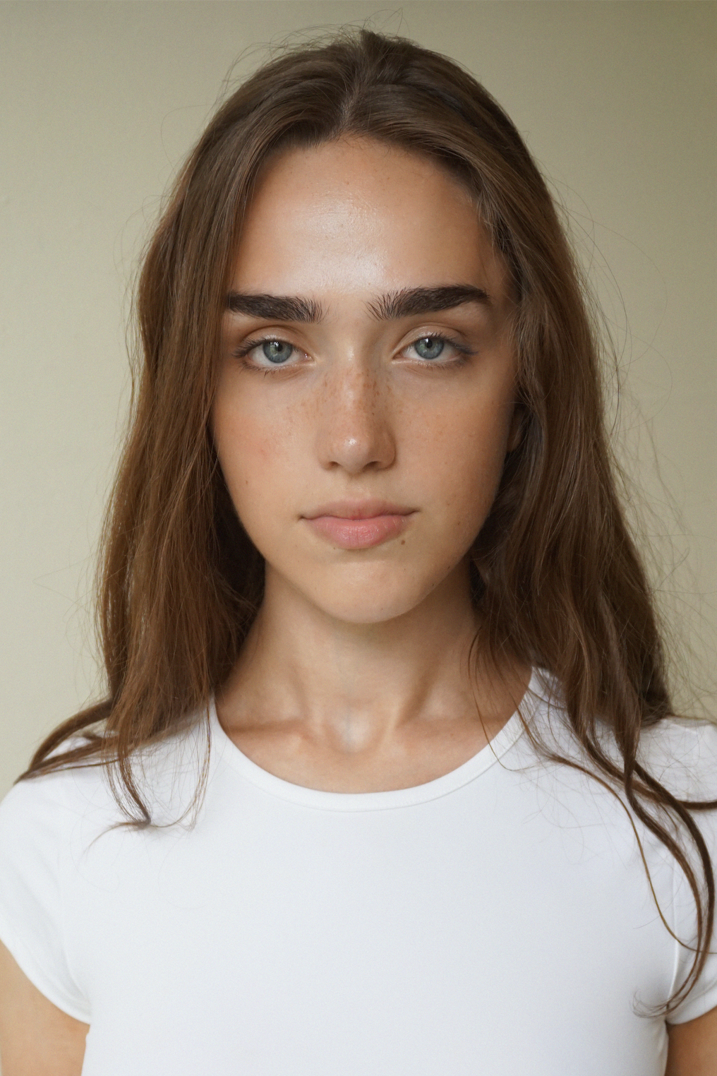 Close-up portrait of a young woman with long, wavy brown hair, blue eyes, freckles, and wearing a white t-shirt, against a plain background.