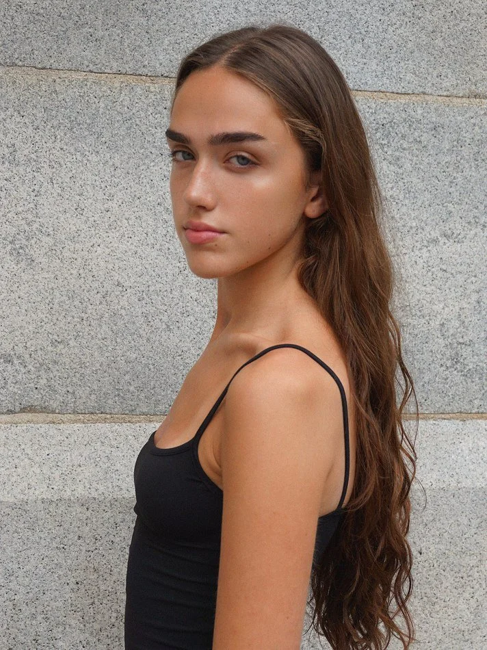 A young woman with long brown hair, wearing a black spaghetti strap top, standing against a light gray stone wall.
