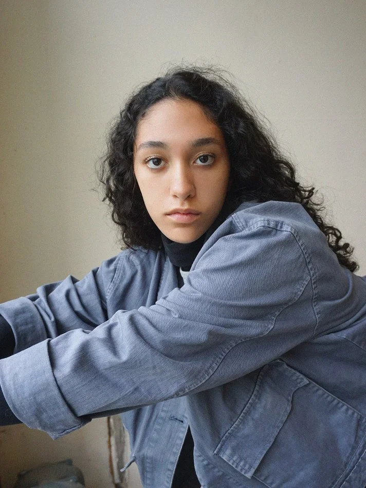 A young woman with curly dark hair and light brown skin looking directly at the camera, wearing a gray denim jacket over a dark shirt, indoors against a neutral wall.