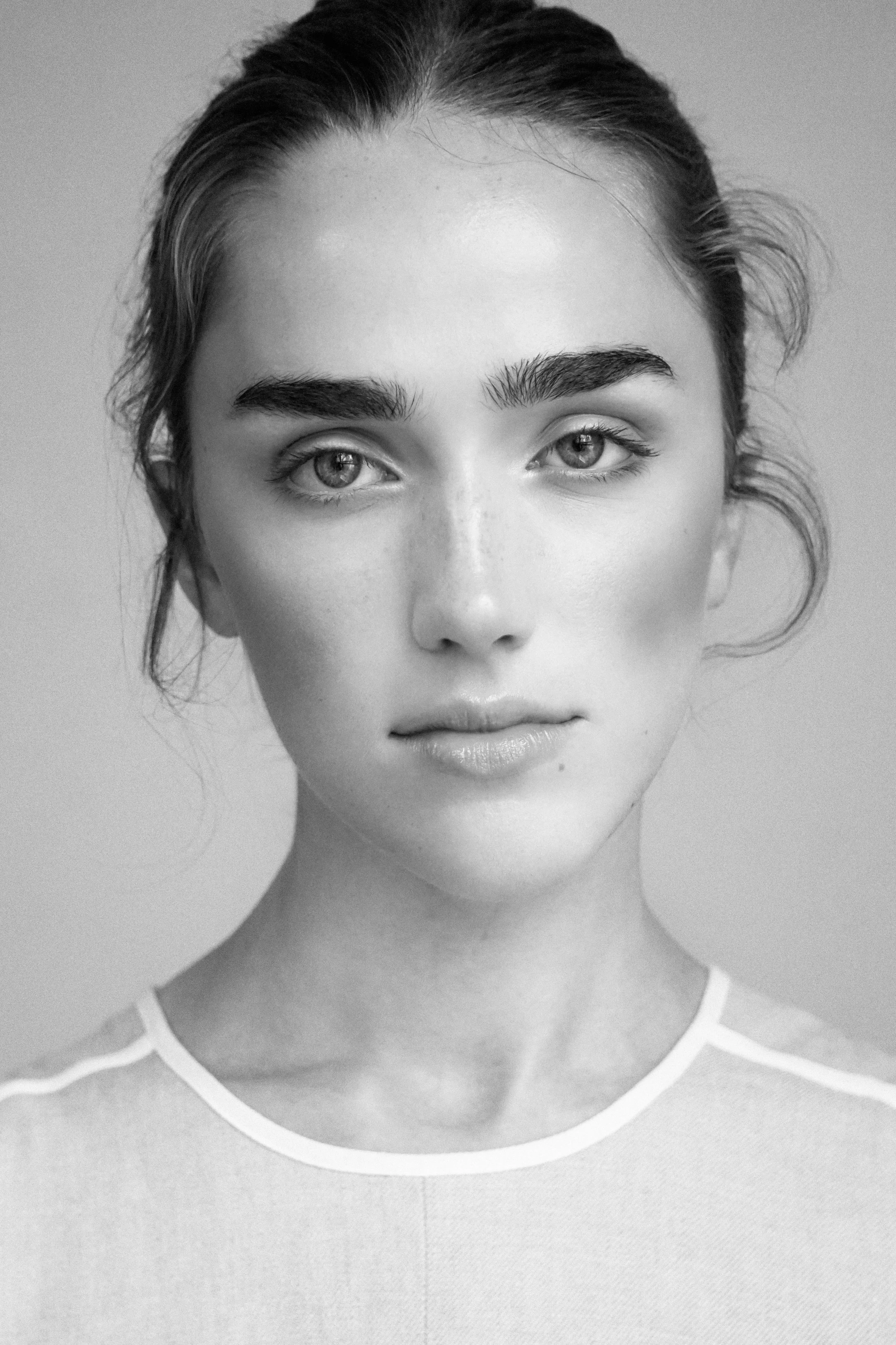 Close-up black and white photograph of a young woman's face, showing her eyes, eyebrows, and part of her lips, with her hair pulled back and a striped shirt visible at the bottom.