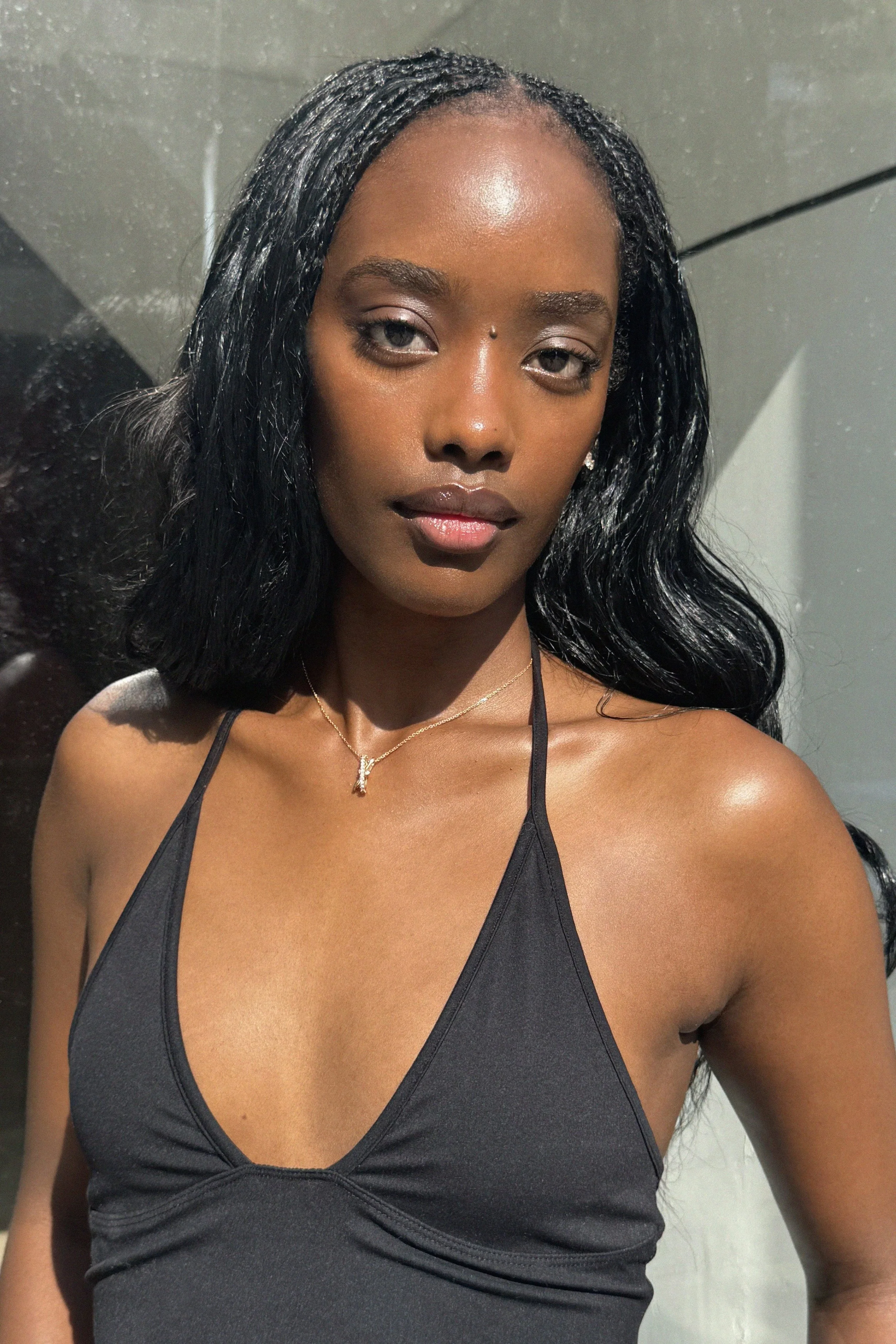 A woman with long black wavy hair, wearing a black spaghetti strap top, a delicate gold necklace, and makeup with glossy lips, standing against a plain wall.