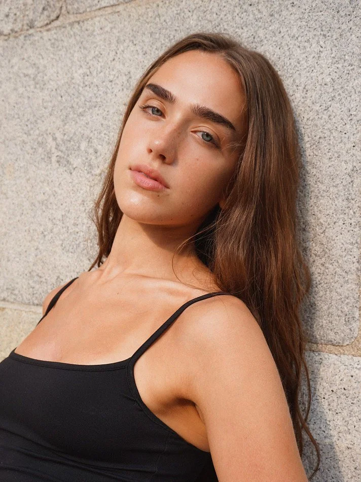 A young woman with long brown hair, light skin, and blue eyes leaning against a stone wall outdoors, wearing a black spaghetti strap top.