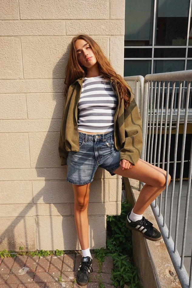 Young woman with long brown hair, wearing a striped shirt, denim shorts, and sneakers, standing outdoors against a beige brick wall with a metal railing nearby.