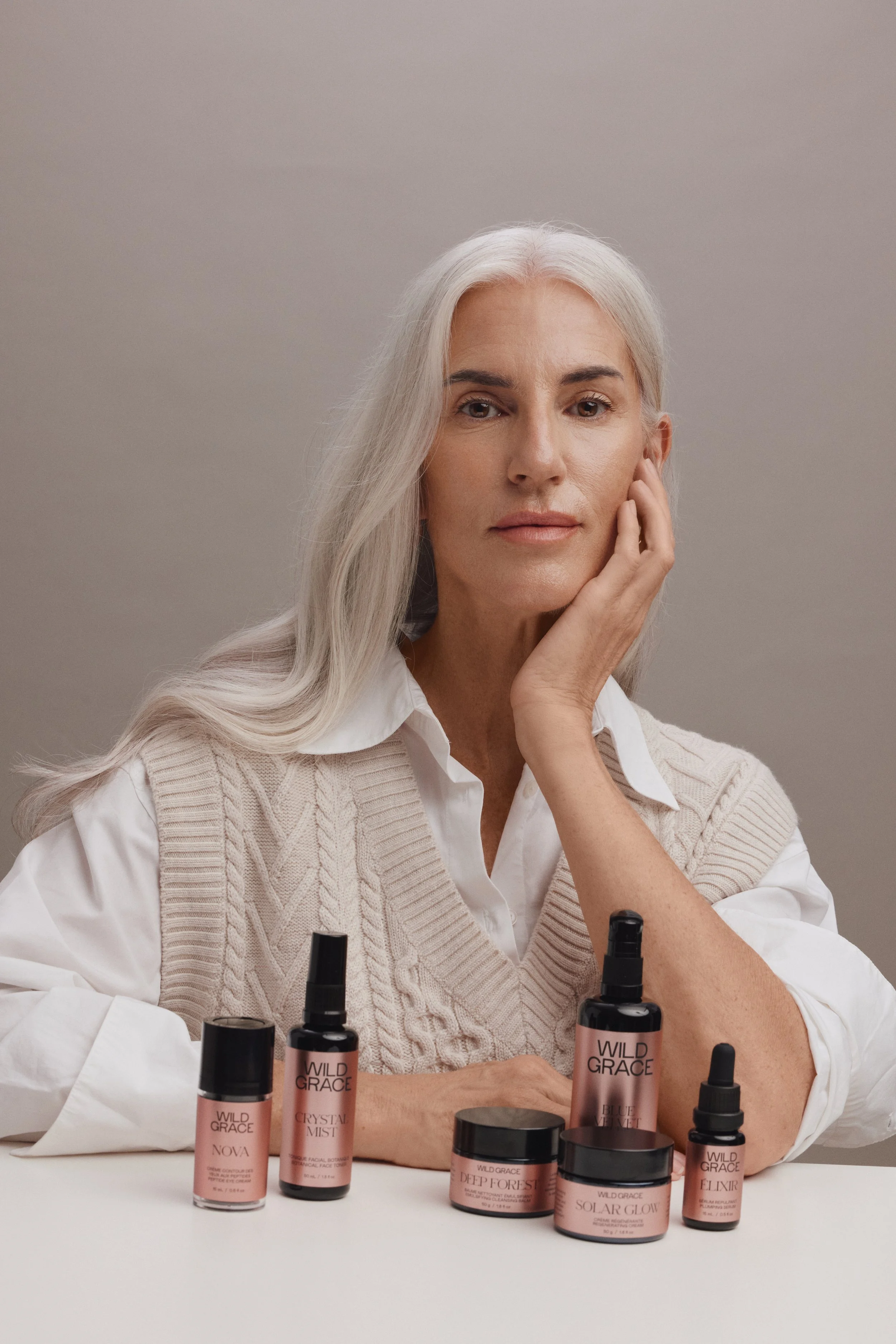 A woman with long gray hair resting her face gently on her hand, sitting behind a table with beauty skincare products from the brand 'Wild Grace' displayed in front of her.