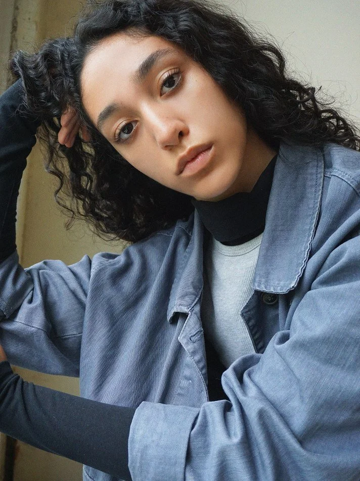 Close-up of a young woman with curly black hair, wearing a gray jacket over a gray shirt, posing with her head resting on her hand.