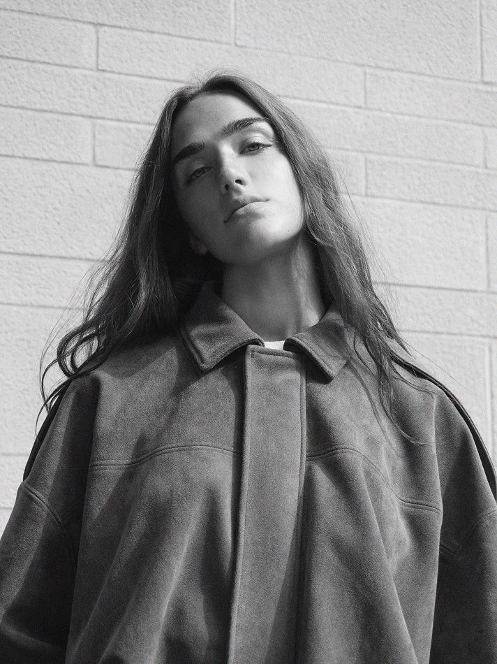 A young woman with long, wavy hair, wearing a large, oversized jacket standing against a brick wall.