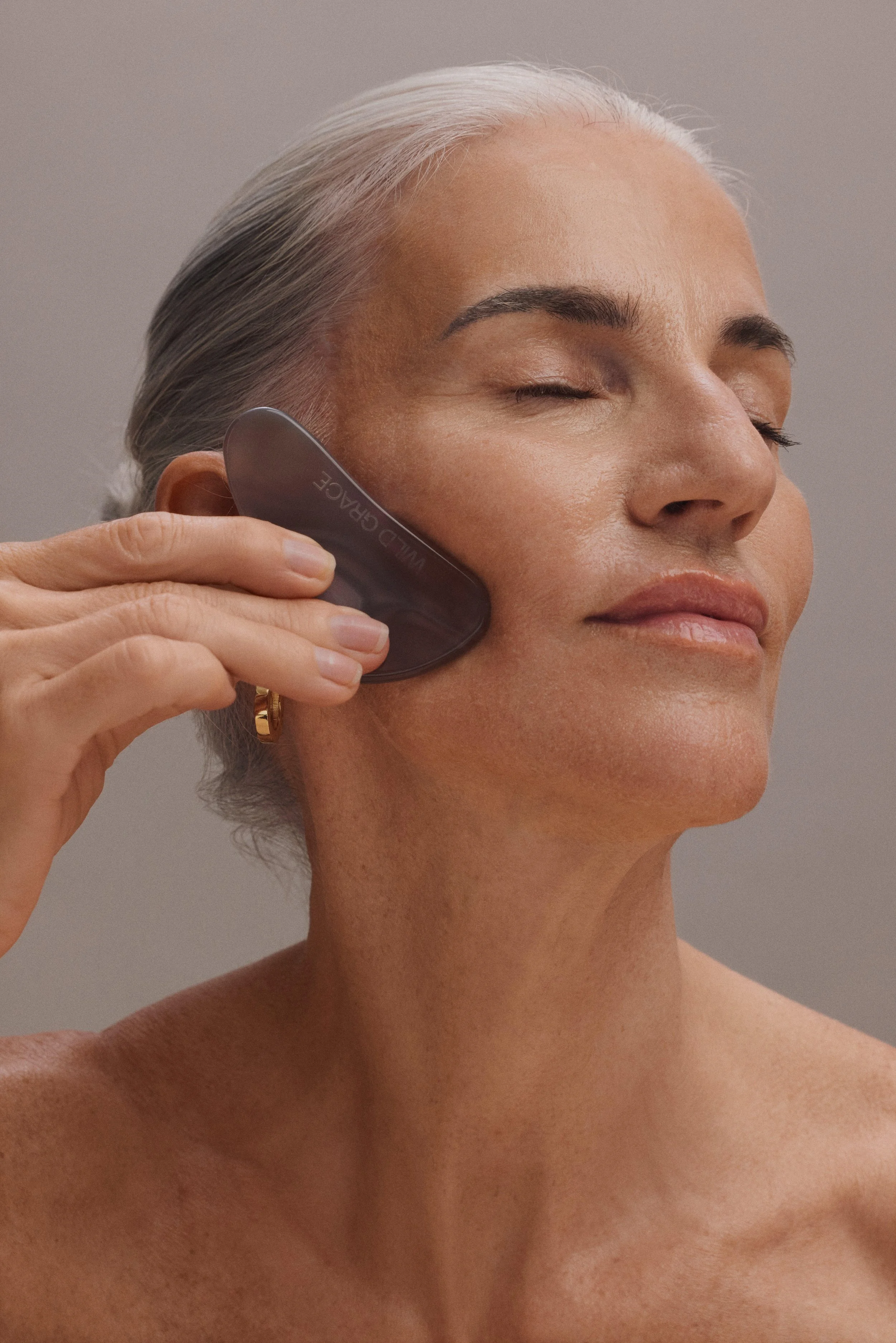A woman with gray hair using a dark colored scalp massager on her scalp, eyes closed, with a neutral facial expression.
