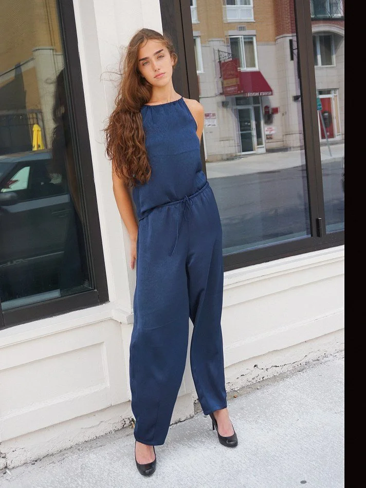 A young woman with long, wavy brown hair standing outside near a large glass window, wearing a blue sleeveless jumpsuit and black high heels.