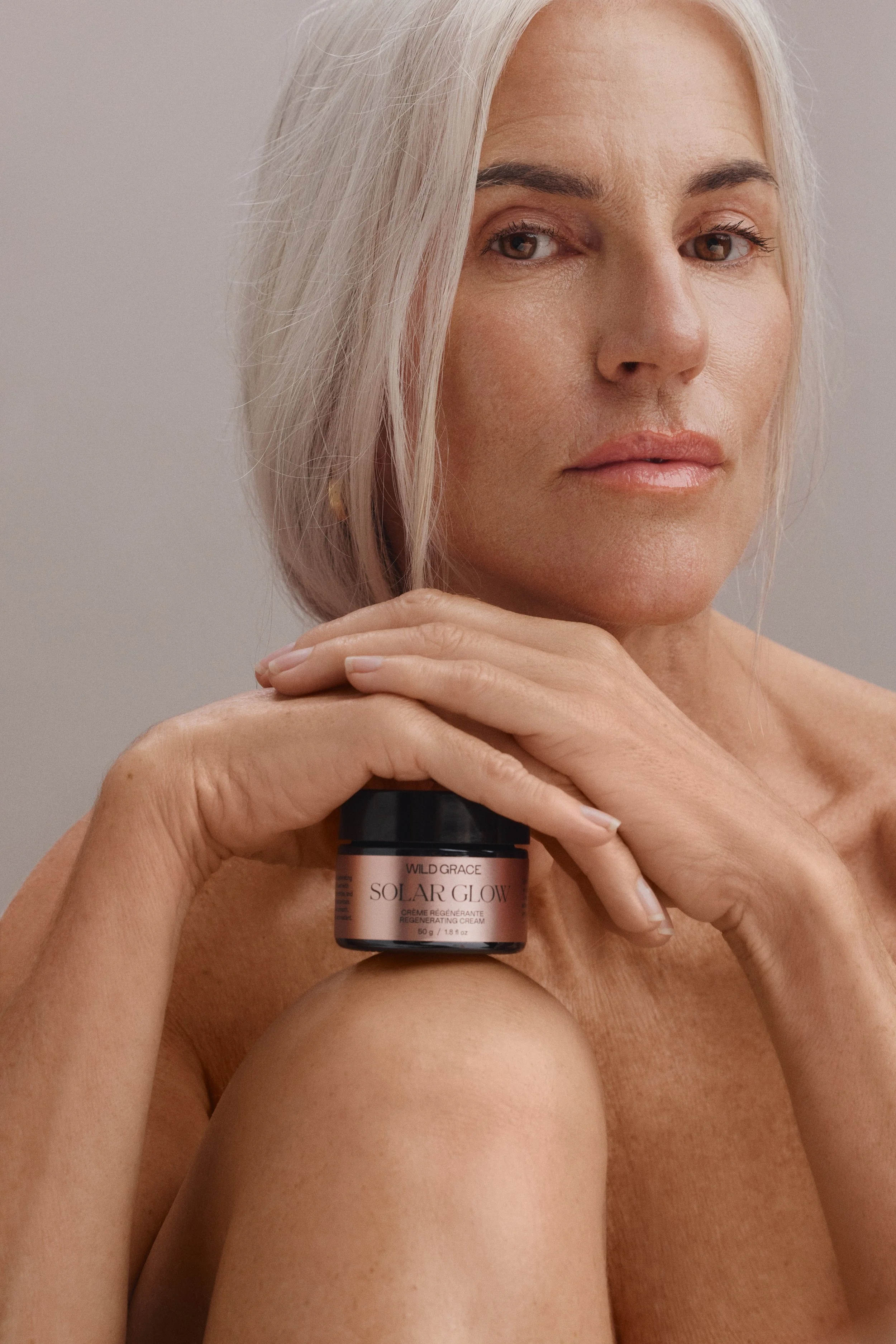 A woman with short blonde hair looking at the camera, holding a jar of Wild Grace Solar Glow regenerating cream near her face, with her arm resting on her shoulder, against a neutral background.