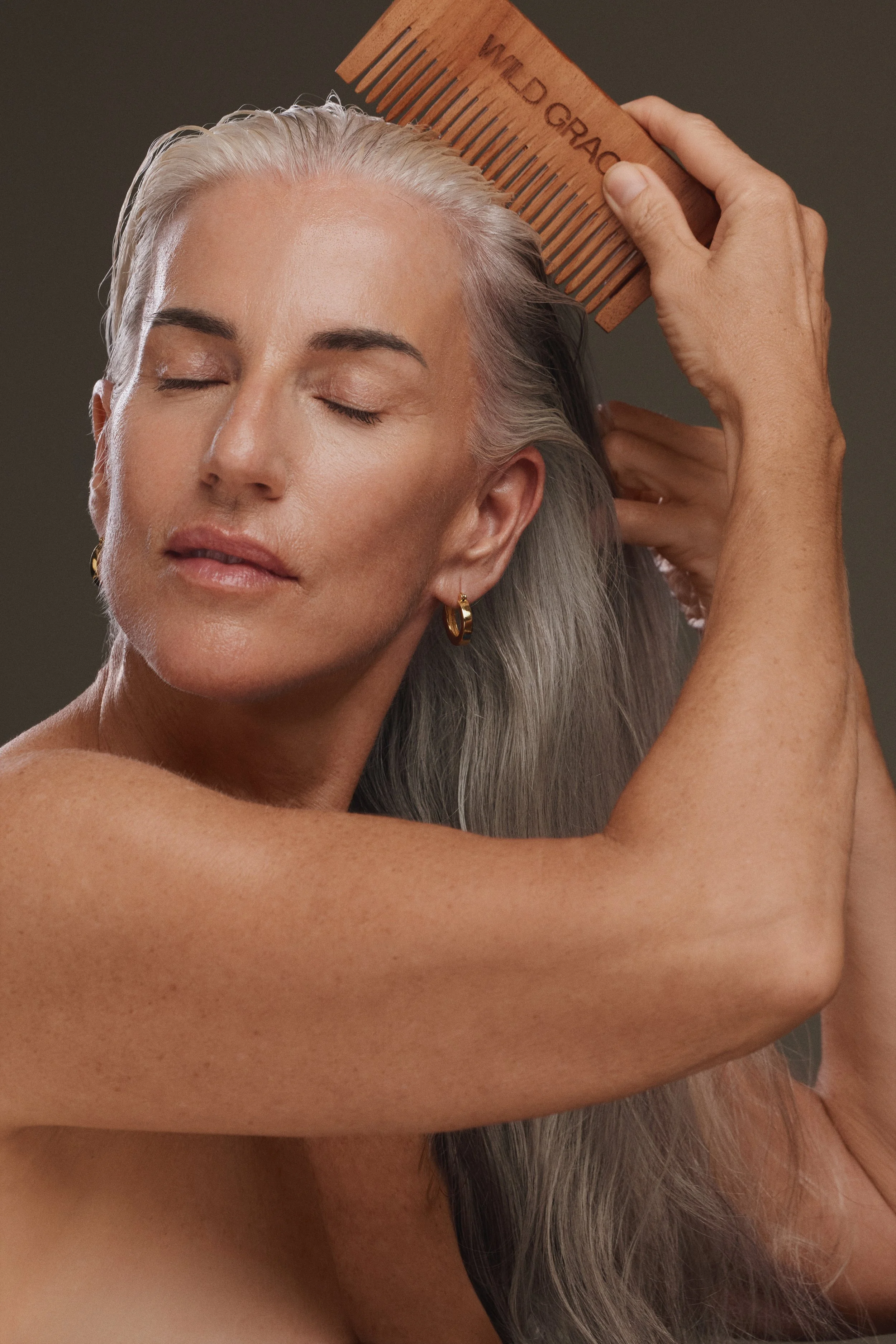 A woman with long gray hair and earrings is combing her hair with a wooden comb that has the word 'WILDGRASS' engraved on it, with her eyes closed and a neutral expression.
