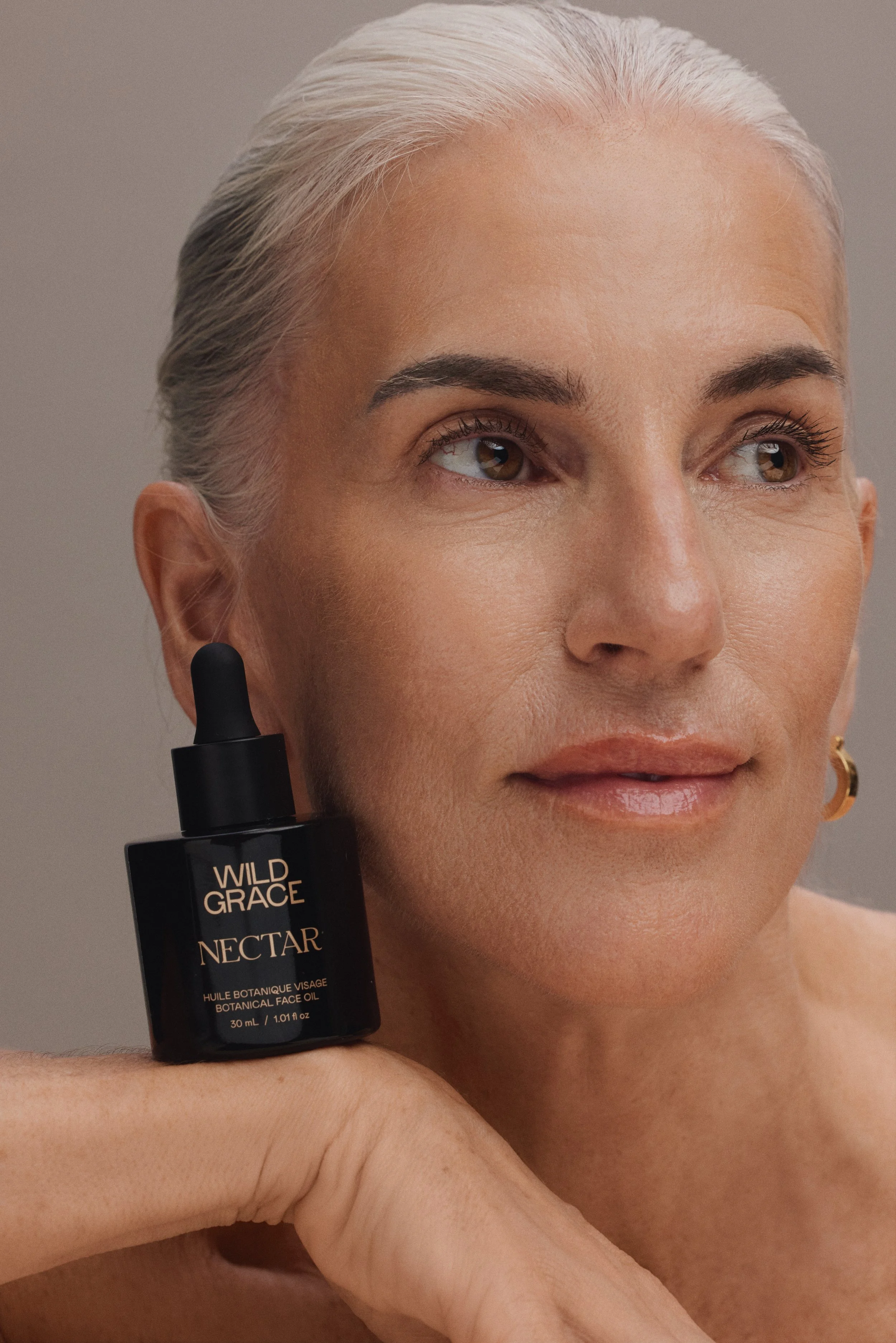 Close-up of a woman with light skin and gray hair, holding a bottle of Wild Grace botanical face oil near her face. She has a neutral expression and is looking slightly to the side against a plain background.