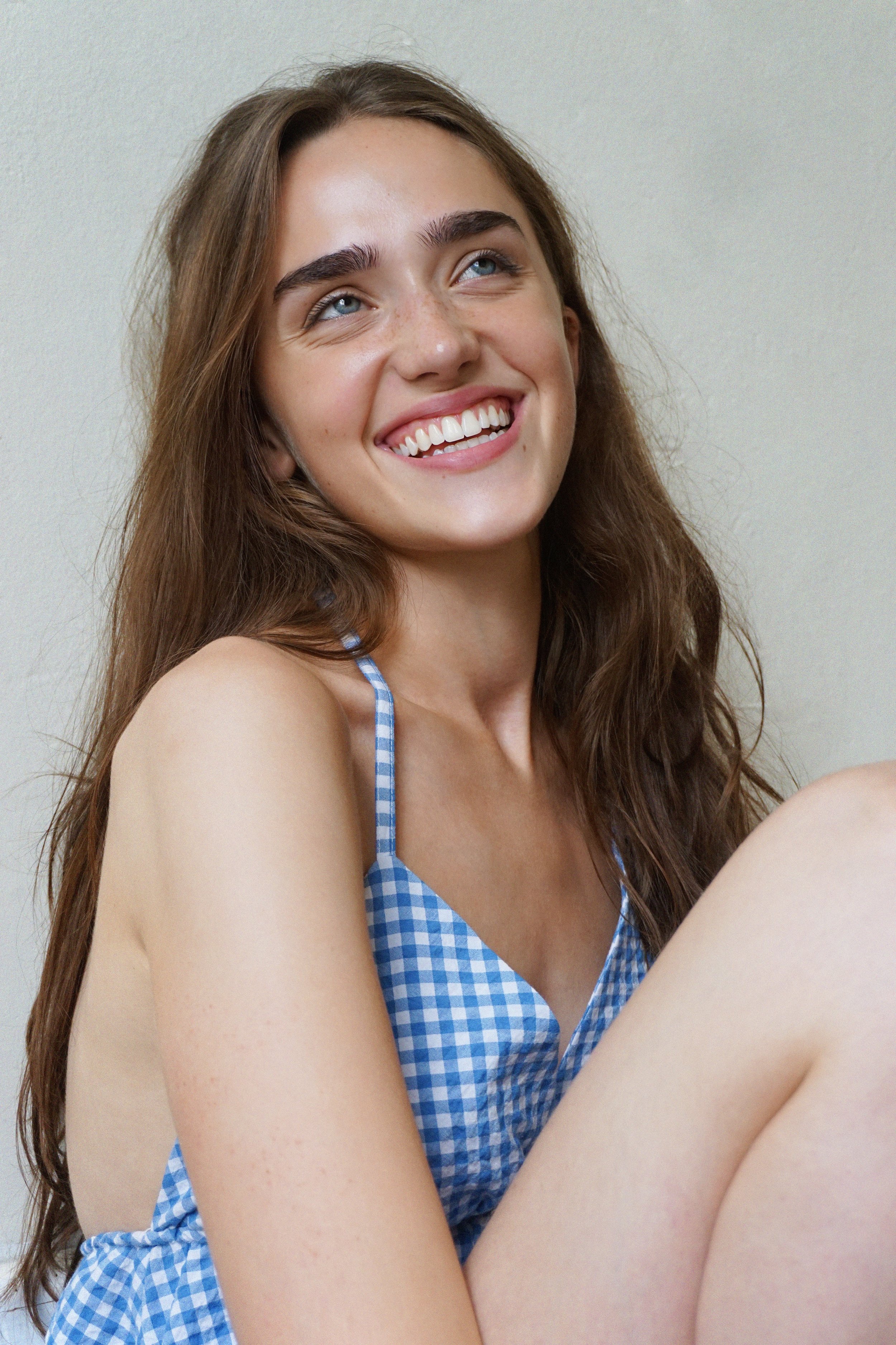 A young woman with long brown hair, blue eyes, and a big smile showing white teeth. She is wearing a blue and white checkered dress with thin straps and is sitting against a plain light-colored background.