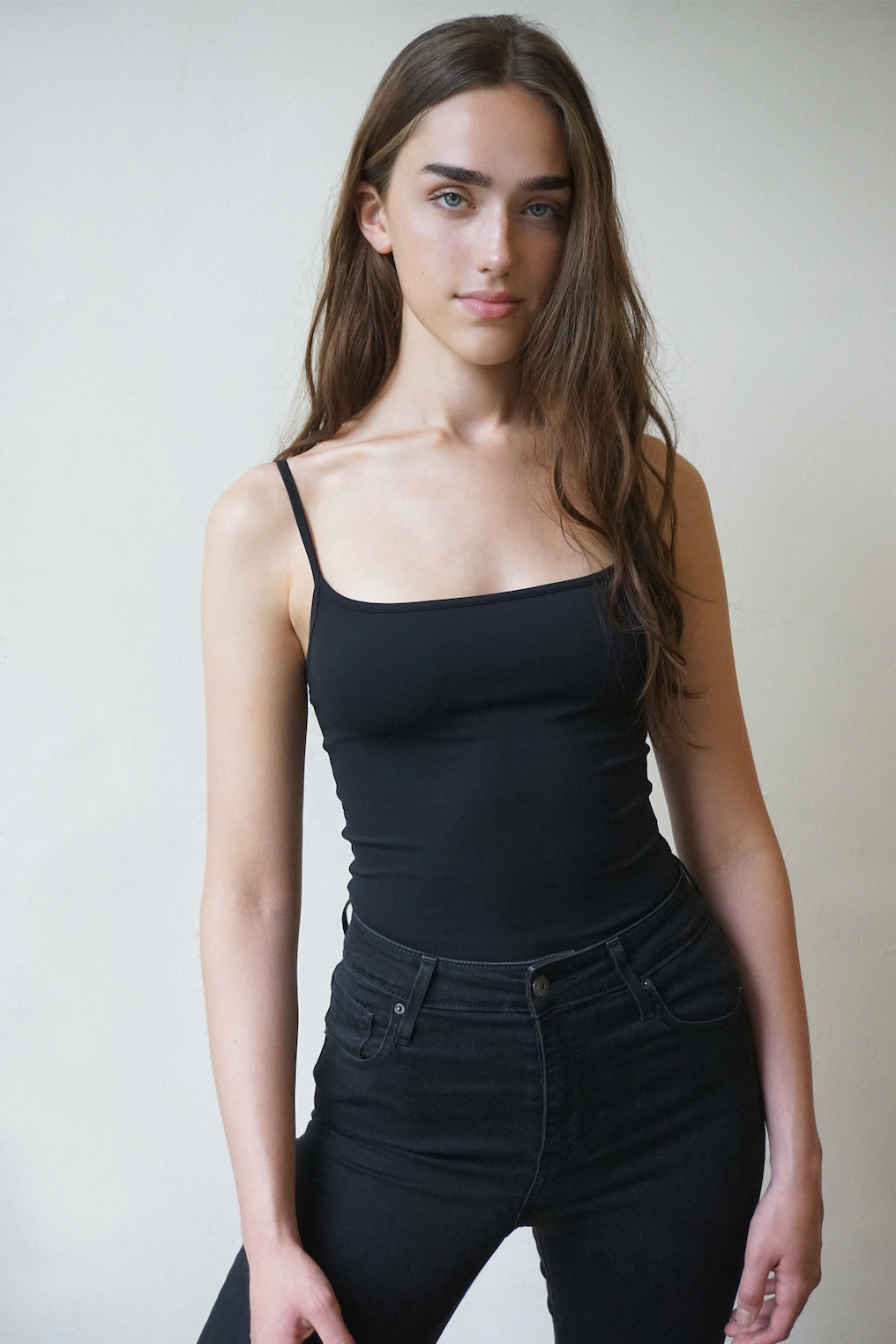 A young woman with long brown hair and blue eyes wearing a black spaghetti strap top and black jeans standing against a plain light-colored wall.