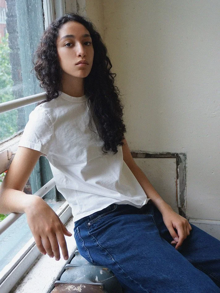 Young woman with curly hair wearing a white t-shirt and blue jeans, sitting on a windowsill inside a room with a plain wall.