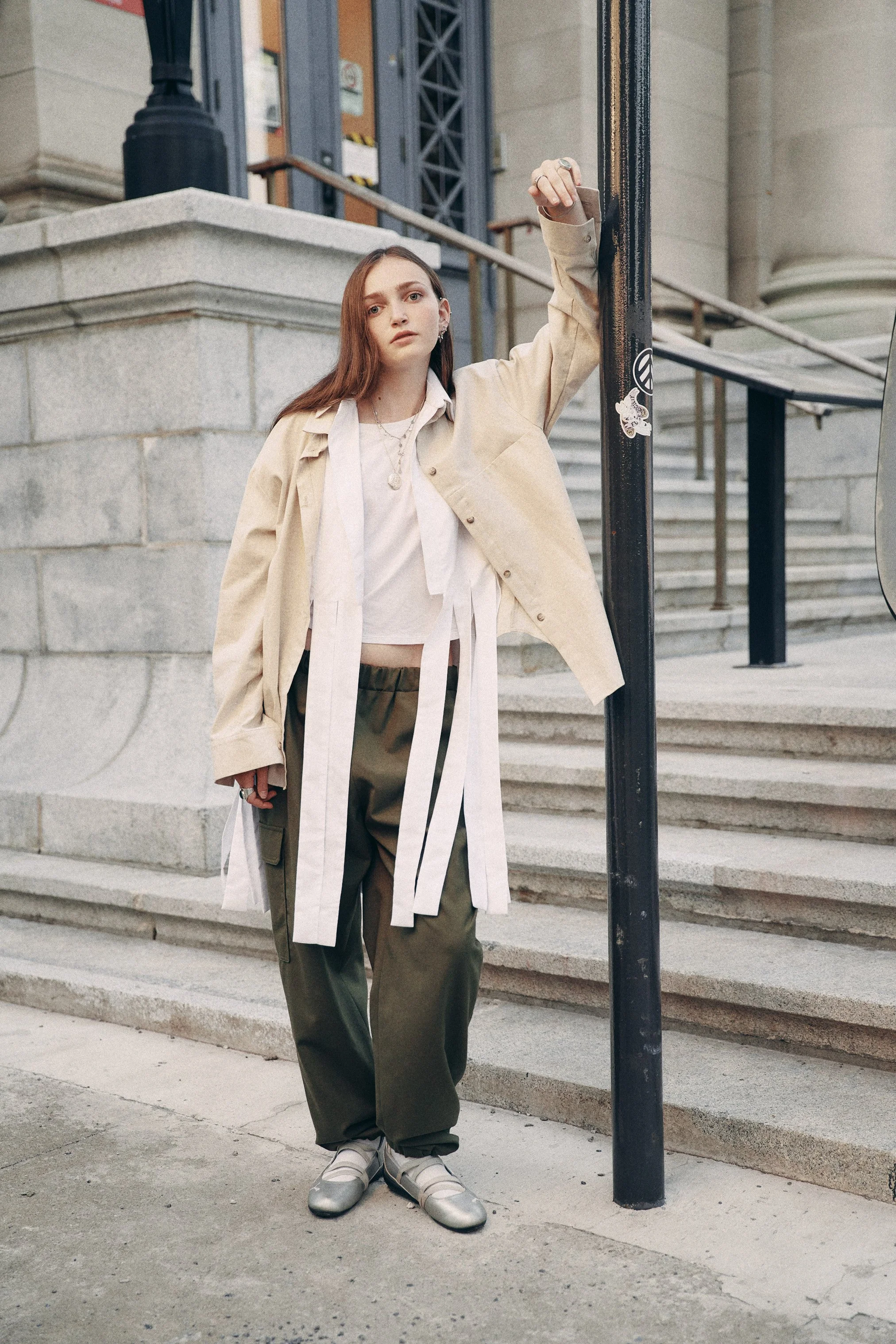 A young woman with long brown hair standing on a city sidewalk near a black lamppost. She is wearing a beige oversized jacket, a white blouse, baggy green cargo pants, and white sneakers. She is leaning against the lamppost with her left arm raised, 