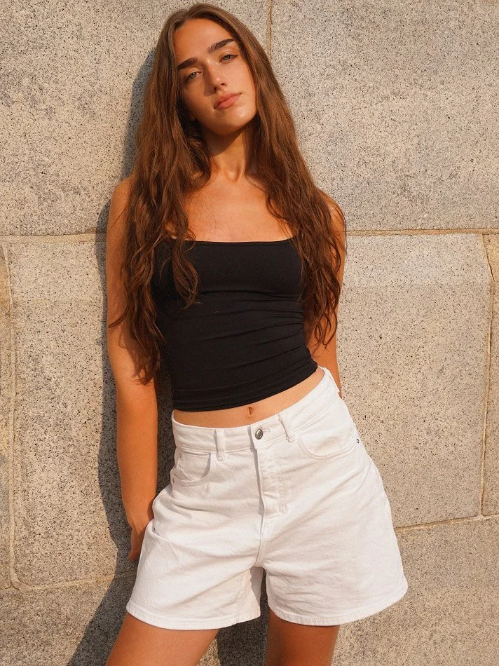 Young woman with long, wavy brown hair leaning against a stone wall, wearing a black tank top and white shorts, looking at the camera.