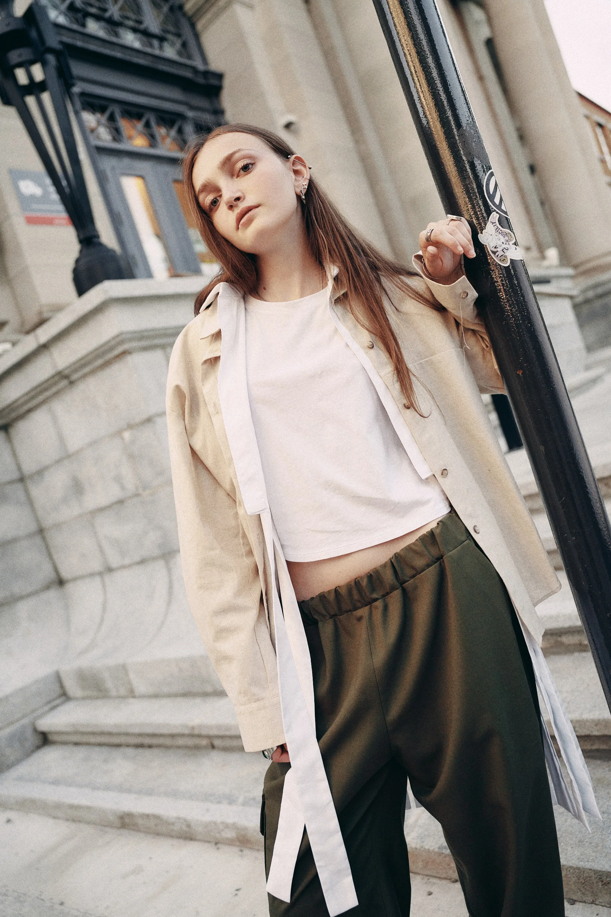 A young woman with long brown hair leaning against a black metal railing outdoors. She is wearing a light-colored oversized shirt over a white crop top and dark green pants. She has earrings and rings and appears to be in an urban setting with stone 