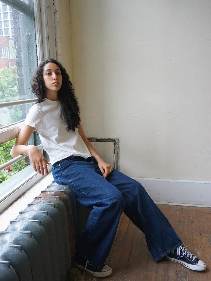A young woman with curly dark hair sitting on a windowsill inside a room, wearing a white t-shirt, blue jeans, and black sneakers, looking at the camera.
