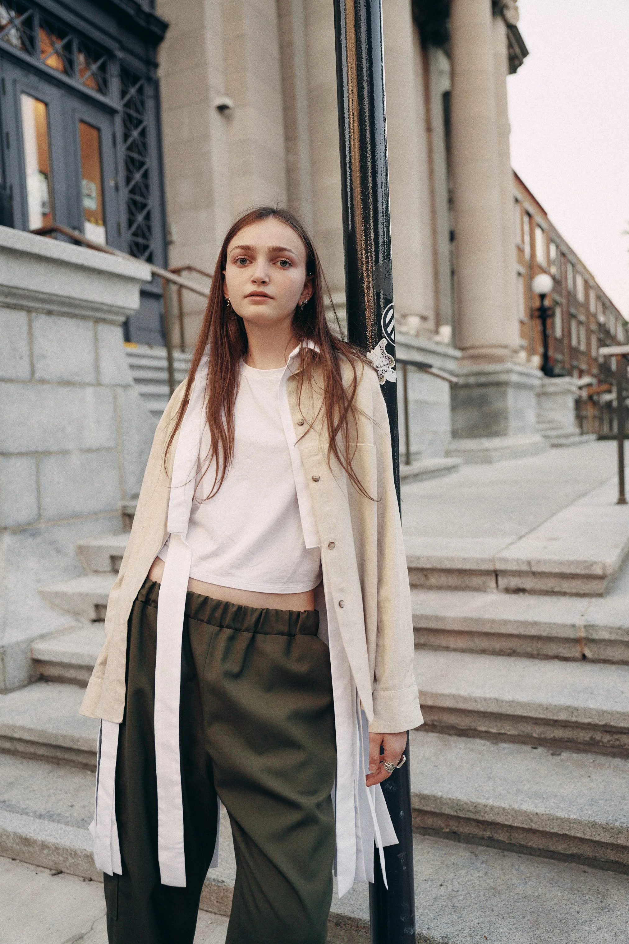 Young woman with long brown hair wearing a beige coat and dark green pants standing on steps in front of a building with columns, holding onto a black street pole.