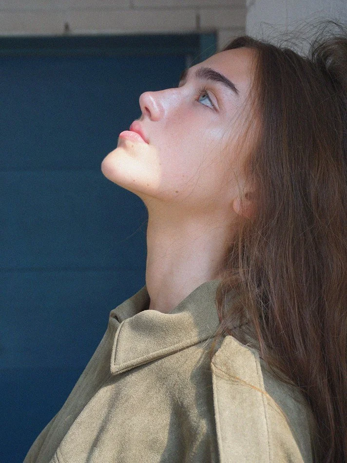 Side profile of a young woman with long brown hair, looking upwards with a neutral expression, wearing a beige coat.