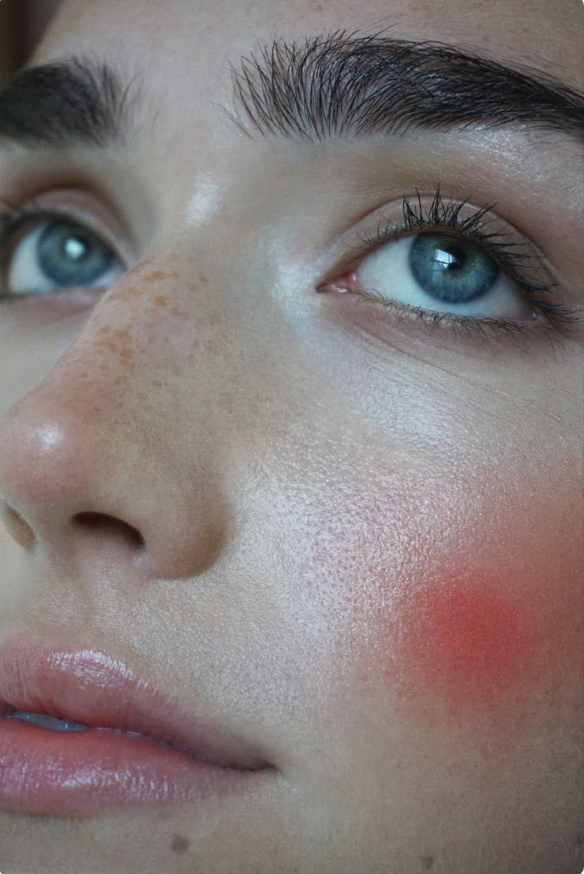 Close-up of a woman's face, showing her blue eyes, dark eyebrows, freckles, and pink blush on her cheeks.